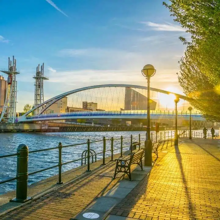 A riverside walkway with benches and trees at sunset, overlooking a modern arched bridge and city buildings—reminiscent of the tranquil beauty offered by Maldives Holiday Packages—with warm sunlight casting long shadows on the pavement.